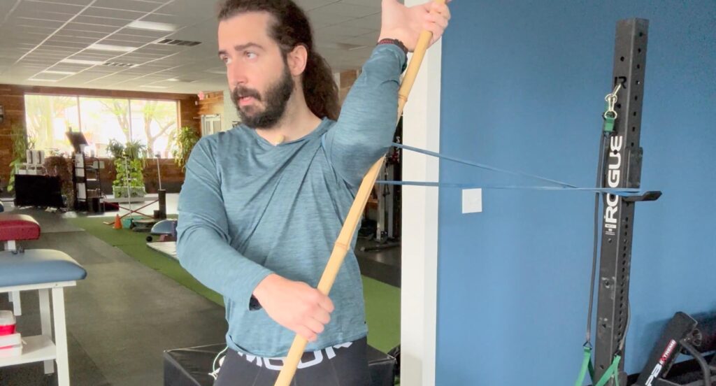 Dr John using a dowel to open up the mobility in his shoulder while it's being pulled back with a band to improve his external rotation for his front rack